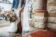 Person in a white dress with brown boots holding a bouquet of flowers, standing next to a stone building.
