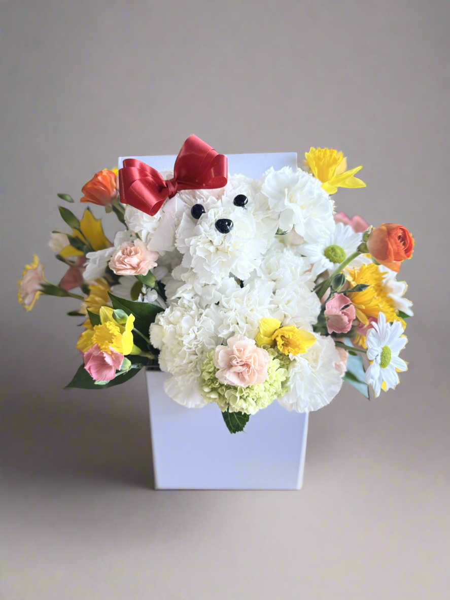 A floral arrangement featuring a mix of fresh flowers including white carnations, daisies, spray roses, and ranunculus, with assorted greenery, presented in a white box with a decorative red bow.