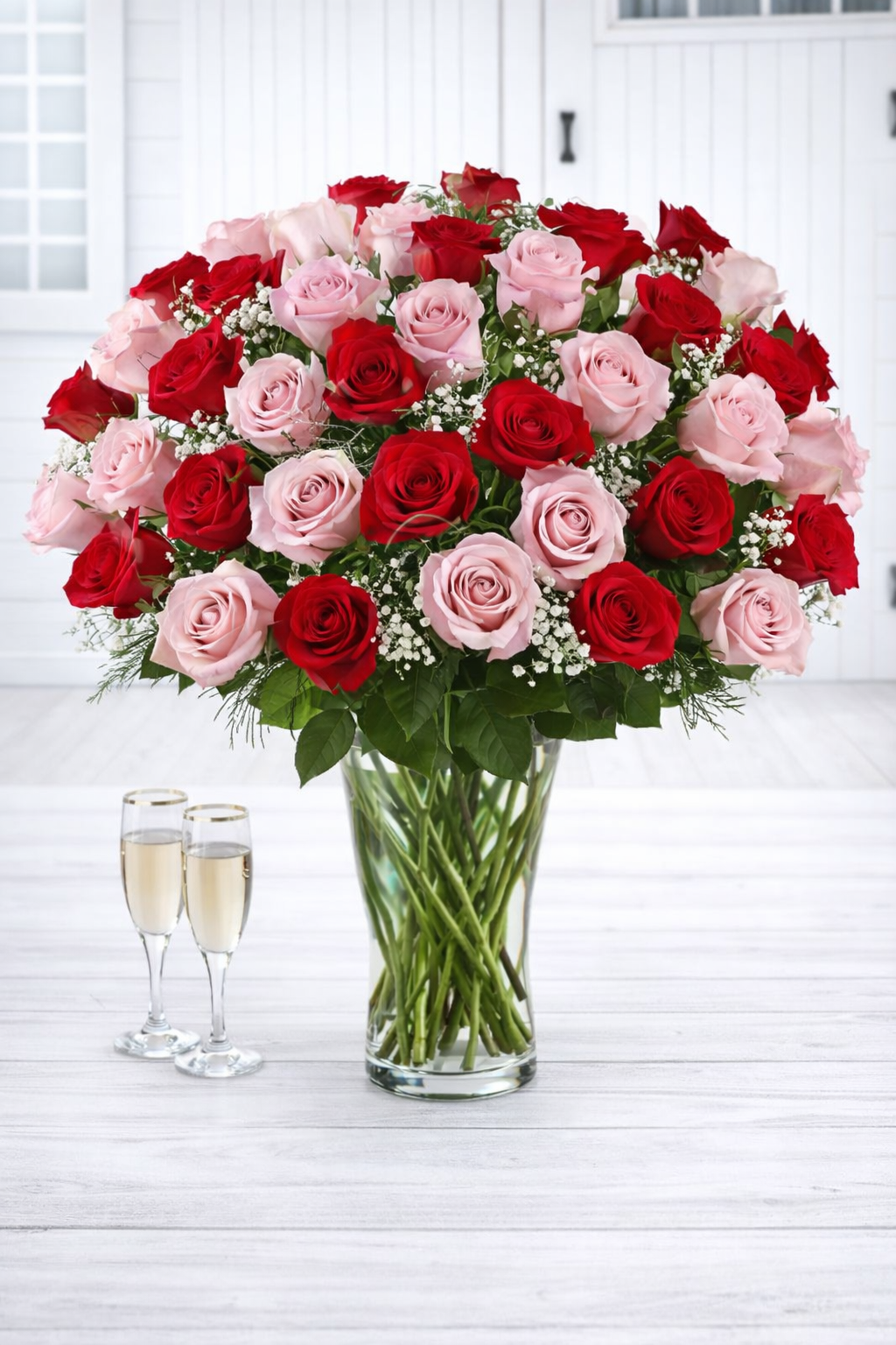 Bouquet of red and pink roses in a clear vase on a white surface with two champagne glasses.